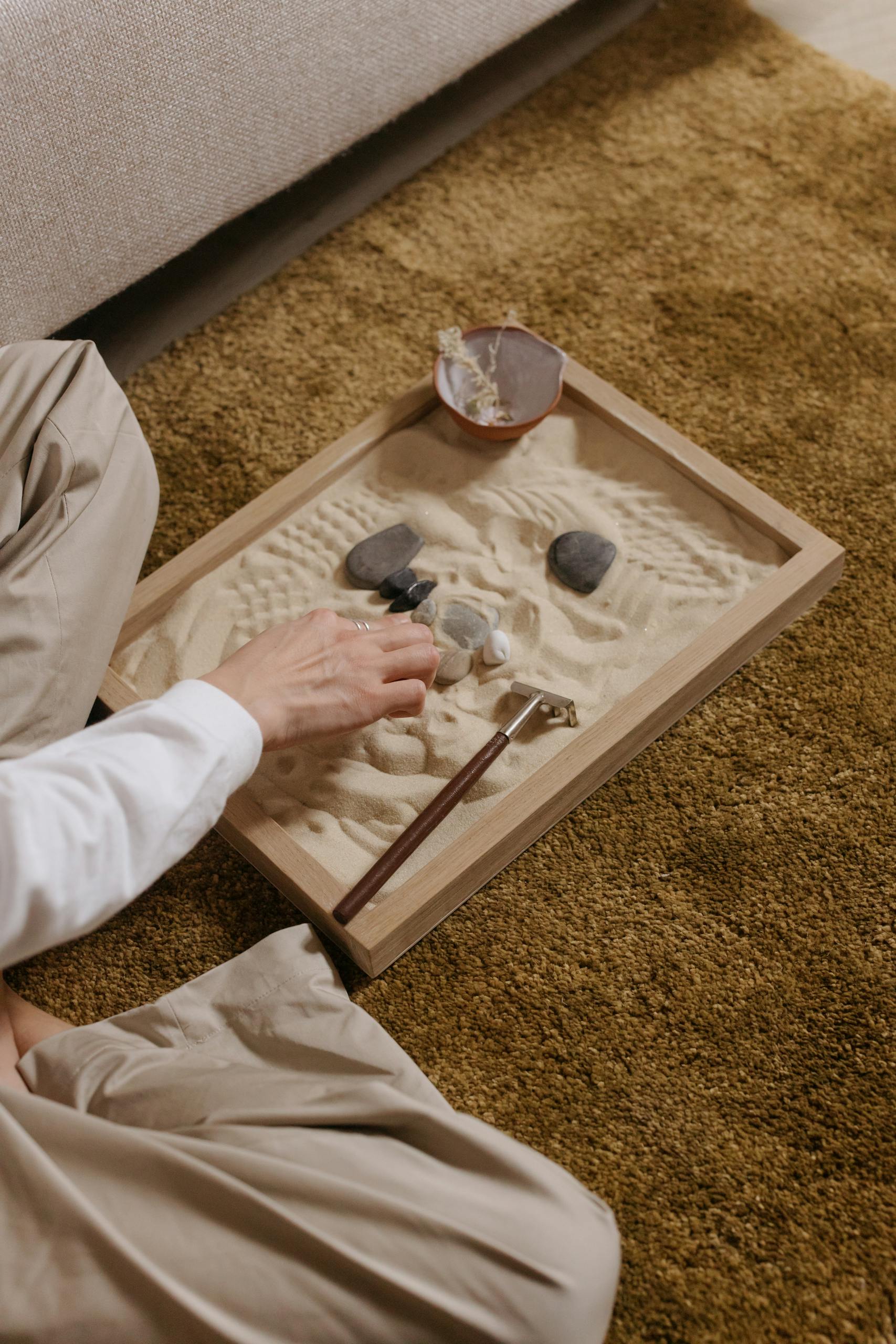 A person creating patterns in a tabletop sand garden with rocks and a rake, promoting relaxation.